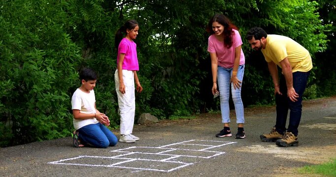 Indian family playing Tikkar Billa, a traditional hopscotch game in home garden, spending quality fun time together with children hopping on squares and enjoying nostalgic outdoor sports activity