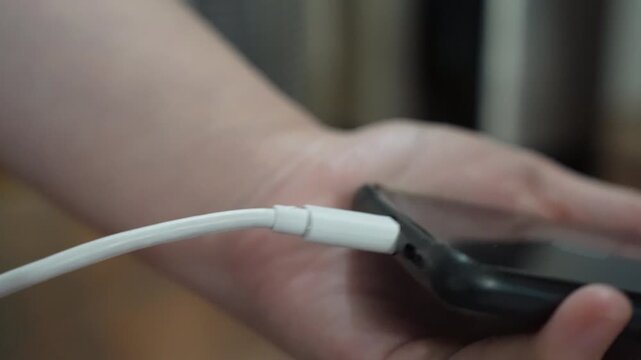Unrecognizable woman lightning cable into smartphone connector to charge sitting at home table. Close-up of the charging connection to smartphone. Phone charging port.