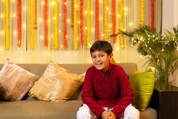 Indian happy handsome little boy looking at the camera wearing traditional kurta sitting on cozy sofa on the occasion of Diwali season in colorful decorated home background - Diwali holidays festive