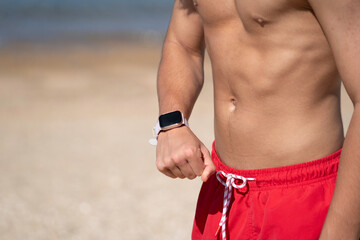 Close up of muscular man checking his smartwatch stands on a beach. Fitness and technology integration concept