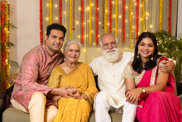 Indian happy family in traditional wear sitting on sofa indoor with smile on the occasion of Diwali season in colorful decorated home background - Diwali holidays festive