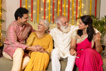 Indian happy family in traditional wear sitting on sofa indoor with smile on the occasion of Diwali season in colorful decorated home background - Diwali holidays festive
