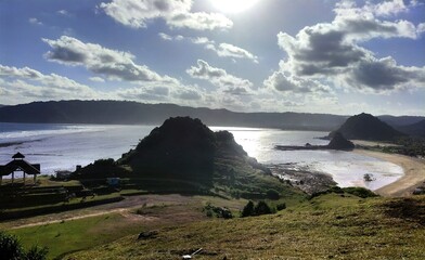 Lombok-Kuta Mandalika, June 11 2025 ; Low tide reveals rocky coastal terrain and tidal pools, with silhouettes of people exploring the shoreline under a bright afternoon sky.