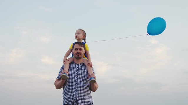 little child girl with colorful balloons rides her father. kid sits shoulders his father against sky. festive mood family holiday outdoors. happy family park. concept children dream fun day teamwork
