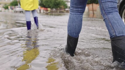 girls in raincoats and rubber boots walk along road flooded with torrential rains, their feet walk through puddles city, splashing water to the sides, the flood is on street, car is driving on water.