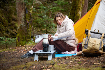 Fototapeta premium Hiker woman preparing coffee outdoor near the camping tent. A female traveler in the forest in early spring holds hot coffee in a tourist mug.