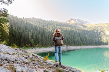 A back view of a female hiker in a hat and with a backpack sitting on a rock overlooking a high mountain lake. Young woman feels freedom and enjoys mountain landscape. Hiking and nature concept.
