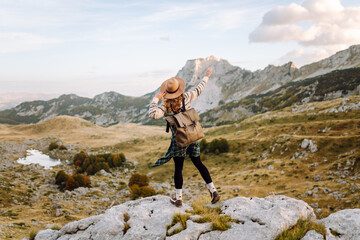 Obraz premium Happy female hiker in hat and authentic sweater with backpack enjoying mountain landscape on mountain trail. Young female hiker feels freedom surrounded by incredible landscapes. Active lifestyle.