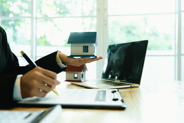 Fototapeta premium A real estate agent holds a house model and signs documents, symbolizing property sale, home purchase, financial agreements, mortgage loans, investment, and ownership in housing deals.