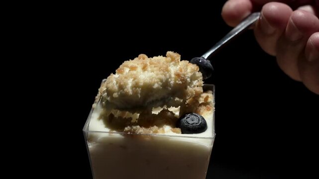 A spoon lifts a creamy layered dessert topped with crumbles and blueberries under dramatic lighting. The texture and rich colors emphasize indulgence and taste.