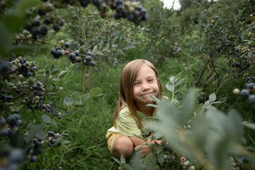 a little girl is peeking out from behind the blueberry bushes and smiling