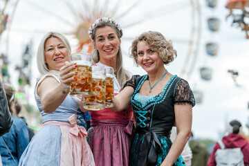 Three joyful women in dirndl outfits toast with beer mugs at an openair folk fest in Bavaria Germany