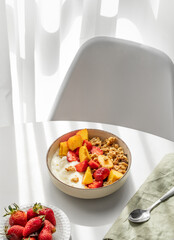 Granola or muesli with natural greek yogurt, strawberry and peach in a bowl on a white table near window