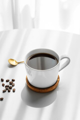 Freshly brewed espresso coffee in a cup on a white table near window with spoon, coffee beans