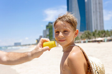 Father, applying sun cream on his  cute blond boy, playing on the sea, hat, sunglasses, swimsuit, sun protection cream, sea and sand background. Concept: love, lifestyle, children, vacations