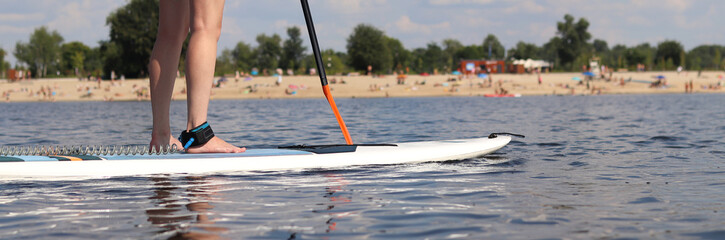 Girl rides a paddle board, close-up of legs. Active summer vacation. Paddle board rental for tourist route, water sports. Girl on a paddle board with a paddle floats on the river near the beach.