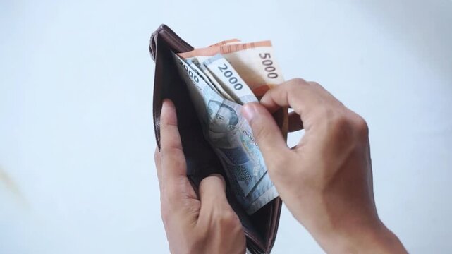 a man's hand is pulling out indonesian money rupiah in a brown leather wallet on a white background
