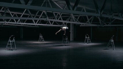 Wide shot of young Caucasian man performing modern dance with stepladder in dark warehouse