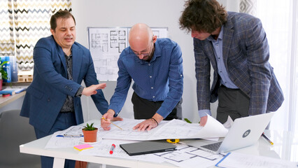 The customer is not satisfied with the construction plan proposed by the architect. A group of men gathered around a table with blueprints and a laptop