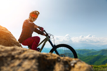 Rider cyclist man wearing mask and full face helmet standing on his bike on top of hill high in mountains. Space copy background for mountain biking