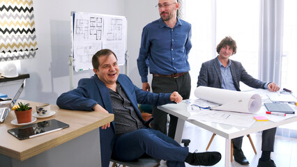 A group of happy entrepreneurs smile and laugh in the conference room after concluding a successful deal. Three men are discussing blueprints