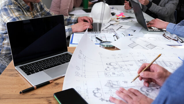Close-up of a civil engineer working on the drawings of a modern business center. A group of people in the office are discussing profitability of project