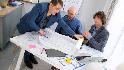 Three men in blue suits are sitting at a table and looking at blueprints. They discuss plans and make sure that everything is in order