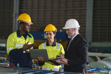 The workers are standing in a factory, looking at a tablet. The engineer talks with the woman's...
