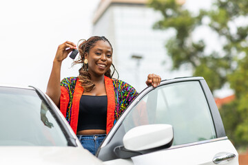 Happy young African woman holding car keys stands by open white car door, smiling.