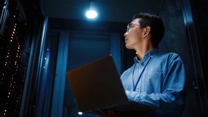 A young Asian male IT engineer works on a laptop in a dimly lit server room with glowing lights from equipment racks overseeing network operations - Powered by Adobe