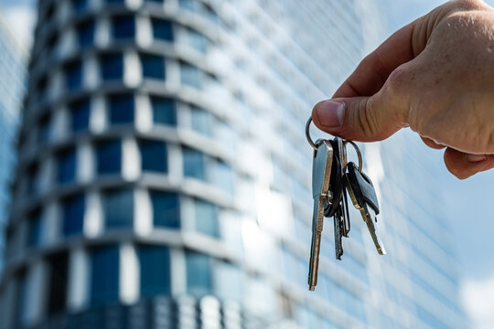 Hand Holding Keys in Front of Modern Glass Office Building. A close-up image of a hand holding a set of metallic keys against the background of a sleek glass skyscraper
