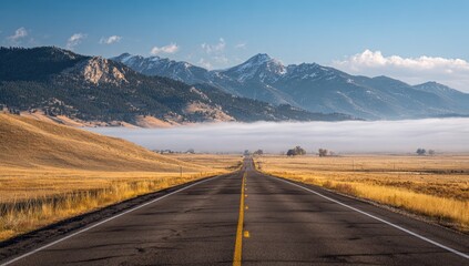 Fototapeta premium A long, straight highway vanishes into a misty valley, framed by autumnal grasslands and snow-capped mountains under a clear, bright sky