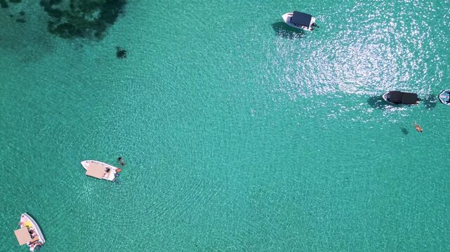 Aerial view of boats anchored in shallow turquoise water near the coast. Clear sea, swimmers, and summer vibes create a relaxing and vibrant beach atmosphere from above