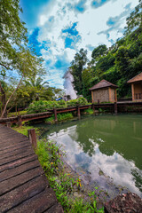 Fototapeta premium Fang hot spring Chiang Mai Thailand. scenic view of a natural hot spring emitting steam in a lush tropical forest. The vibrant greenery, blue sky, and traditional wooden structures in the background