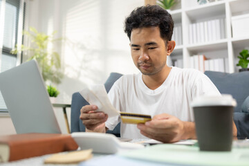 A man is sitting on a couch and looking at a receipt on a laptop. He is holding a credit card and he is checking his bank account. Concept of financial responsibility

