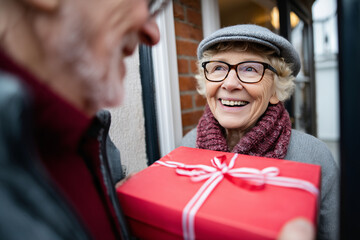 Elderly European couple standing at front door holding gift box, being welcomed by family, 
