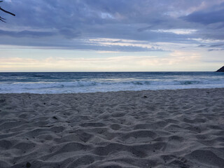 Beach at sunset with sand in the foreground and ocean with waves breaking on the shore – Cloudy sky with pink and blue tones on the horizon