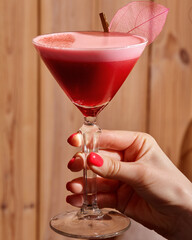 Woman hand holding elegant glass with red layered cocktail decorated with pink leaf
