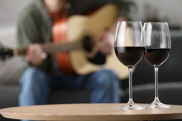 Glasses with red wine on table and man playing guitar indoors, selective focus