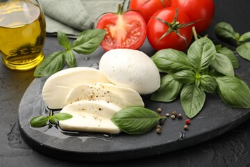 Tasty mozzarella cheese, tomatoes, basil, peppercorns and oil on black table, closeup