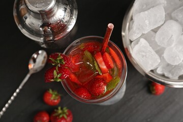 Refreshing strawberry cocktail in glass, metal shaker and ice in bucket on dark table, flat lay
