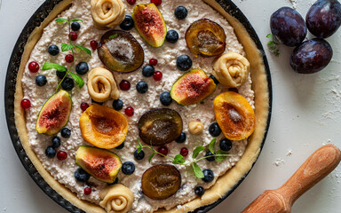 A tart or cottage cheese pie with fruits and berries in a mold is prepared for baking and a rolling pin on a light background or table. View from above.