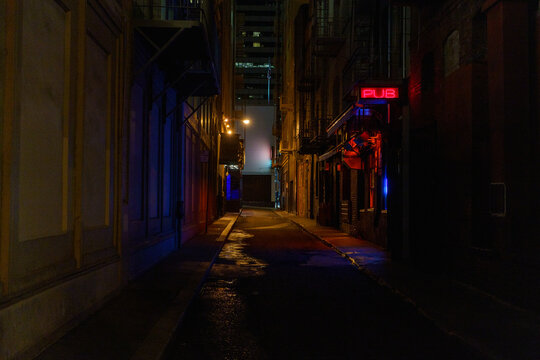 A bright red neon pub sign illuminates a dark, wet city alley, casting colorful reflections onto the pavement.
