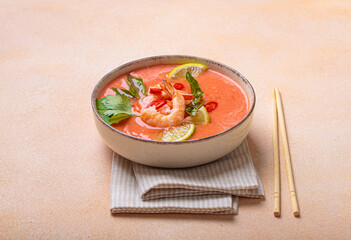 Creamy flavourful tom yum soup, a popular dish of Thailand, is served in a bowl, garnished with fresh shrimp, lime wedges, herbs, and chili peppers, accompanied by chopsticks on a linen napkin