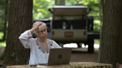 Frustrated, unhappy mature woman sits at a table in a forest campground, using a computer and smartphone while a teardrop camper trailer is visible in the background.
