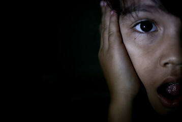 Fototapeta premium Portrait in the dark of a scared and upset 5 year old girl hand on mouth on dark background in profile