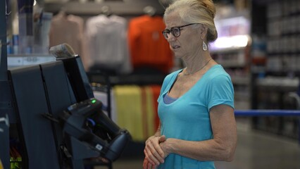 A mature woman stands at a self checkout kiosk in a hardware store, holding her credit card while preparing to complete her purchase.