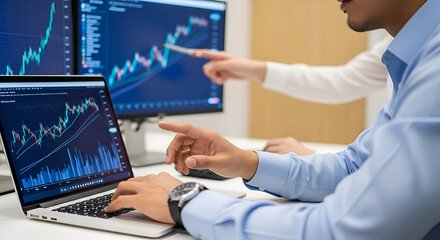 Man analyzing stock market data on laptop and monitor with a colleague pointing at the screen