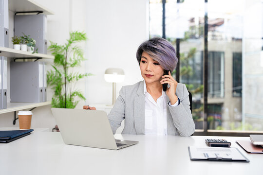 Mature middle aged business woman, 40s professional lady executive manager talking on the phone making business call on cellphone at work in office using laptop computer.