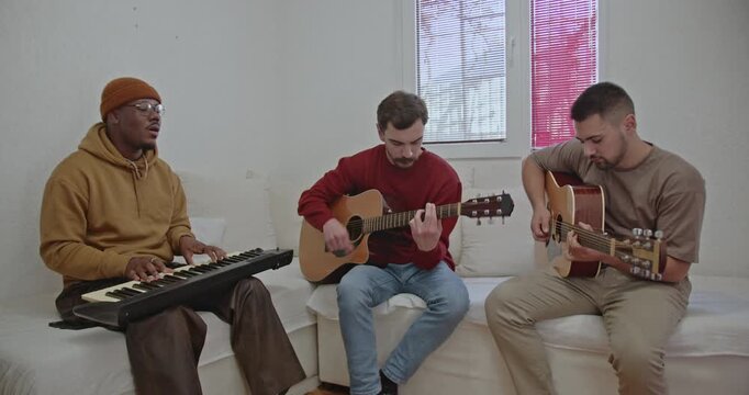 Three friends from around the world are having a jam session in their apartment. The diverse trio consists of a man playing keyboard, and two others on guitars.